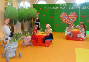 Kilka dziewczynek siedzi na rozstawionych ławeczkach z lalkami, jedna z dziewczynek stoi z boku i trzyma w ręku mikrofon recytuje wiersz. Z lewej strony stoi uśmiechnięta dziewczynka z wózkiem.